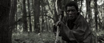 Movie still from “Emancipation” (2022), directed by Antoine Fuqua – A black and white photo of a man holding a stick in the woods; Medium shot, High angle