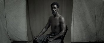 Movie still from “Emancipation” (2022), directed by Antoine Fuqua – A black and white photo of a man sitting in a chair; Medium shot, Low angle