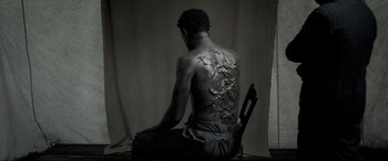 Movie still from “Emancipation” (2022), directed by Antoine Fuqua – A man sitting on a chair with a tattoo on his back; Medium shot, Overhead angle