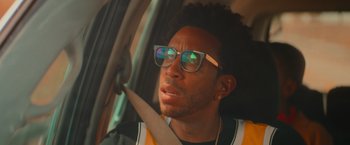 Movie still from “End of the Road” (2022), directed by Millicent Shelton – A young man driving in a car wearing glasses; Close Up shot, Low angle