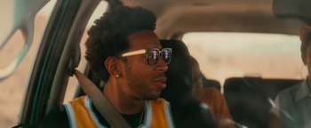 Movie still from “End of the Road” (2022), directed by Millicent Shelton – A man sitting in the back seat of a car wearing sunglasses; Close Up shot, Over the shoulder angle