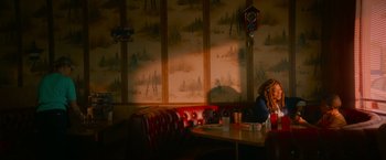 Movie still from “End of the Road” (2022), directed by Millicent Shelton – A room with a table and a clock in the corner of the room; Wide shot, Low angle