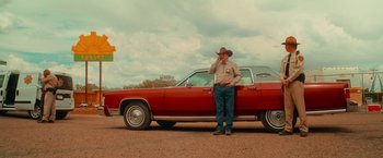 Movie still from “End of the Road” (2022), directed by Millicent Shelton – A man standing next to a red car on a dirt road; Wide shot, Low angle