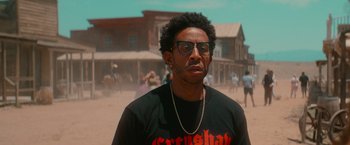 Movie still from “End of the Road” (2022), directed by Millicent Shelton – A man in a black t - shirt is standing in front of some buildings; Close Up shot, Over the shoulder angle