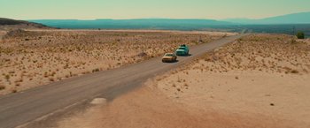 Movie still from “End of the Road” (2022), directed by Millicent Shelton – A couple of cars driving down a dirt road in the middle of the desert; Extreme Wide shot, High angle