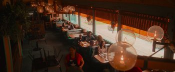 Movie still from “End of the Road” (2022), directed by Millicent Shelton – People are sitting in a restaurant with a view of the outside; Extreme Wide shot, High angle