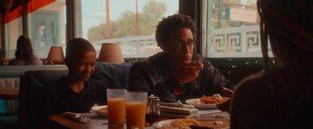 Movie still from “End of the Road” (2022), directed by Millicent Shelton – A man sitting at a table eating a piece of food; Medium shot, Over the shoulder angle