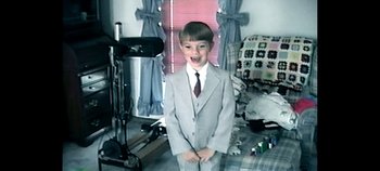 Movie still from “Enemies of the State” (2020), directed by Sonia Kennebeck – A boy in a suit and tie standing in front of a camera; Wide shot, High angle