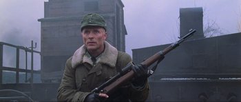 Movie still from “Enemy at the Gates” (2001), directed by Jean-Jacques Annaud – A man holding a rifle while standing in front of a building; Medium shot, Low angle