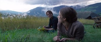 Movie still from “A Hidden Life” (2019), directed by Terrence Malick – Two people sitting in a field with a mountain in the background; Medium shot, Low angle