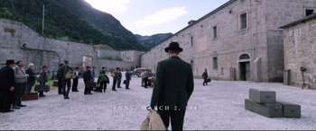 Movie still from “A Hidden Life” (2019), directed by Terrence Malick – A man in a green suit and a black hat is holding a suitcase; Extreme Wide shot, High angle