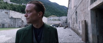 Movie still from “A Hidden Life” (2019), directed by Terrence Malick – A man wearing a green jacket standing in front of a stone wall; Close Up shot, Over the shoulder angle