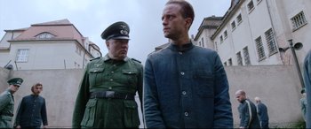 Movie still from “A Hidden Life” (2019), directed by Terrence Malick – A man in a blue jacket stands next to a man in a green uniform; Medium shot, Over the shoulder angle