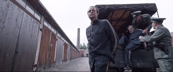 Movie still from “A Hidden Life” (2019), directed by Terrence Malick – A man standing next to a building in a lot; Medium shot, Low angle