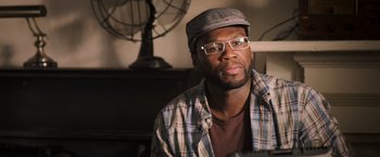 Movie still from “Escape Plan” (2013), directed by Mikael Håfström – A man wearing glasses and a hat sitting in front of a desk; Close Up shot, Over the shoulder angle