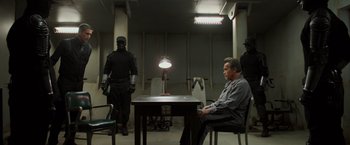 Movie still from “Escape Plan” (2013), directed by Mikael Håfström – A man sitting at a table in front of two other men in a room; Wide shot, Over the shoulder angle