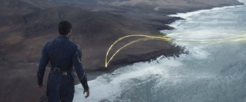 Movie still from “Eternals” (2021), directed by Chloé Zhao – A man flying a kite over a body of water; Extreme Wide shot, Overhead angle