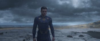 Movie still from “Eternals” (2021), directed by Chloé Zhao – A man standing on top of a beach under a cloudy sky; Medium shot, Low angle