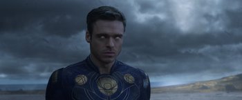 Movie still from “Eternals” (2021), directed by Chloé Zhao – A man in a blue and gold outfit standing in front of a cloudy sky; Close Up shot, Over the shoulder angle