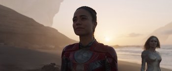 Movie still from “Eternals” (2021), directed by Chloé Zhao – A woman in a red and black costume standing on a beach; Medium shot, Over the shoulder angle