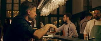 Movie still from “A Taste of Hunger” (2021), directed by Christoffer Boe – A man sitting at a bar holding a glass of whiskey; Medium shot, Over the shoulder angle
