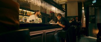 Movie still from “A Taste of Hunger” (2021), directed by Christoffer Boe – A man sitting at a counter in front of a bar; Medium shot, Over the shoulder angle