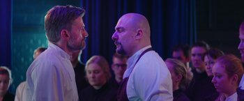 Movie still from “A Taste of Hunger” (2021), directed by Christoffer Boe – Two men are facing each other in front of an audience; Close Up shot, Over the shoulder angle