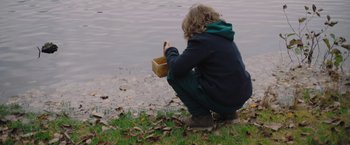 Movie still from “A Taste of Hunger” (2021), directed by Christoffer Boe – A person kneeling on the grass near a body of water; Medium shot, Low angle