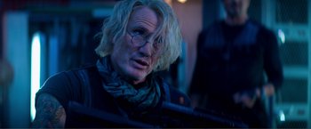 Movie still from “Expend4bles” (2023), directed by Scott Waugh – A man with blonde hair wearing glasses and a scarf; Close Up shot, Low angle