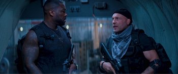 Movie still from “Expend4bles” (2023), directed by Scott Waugh – Two men are standing next to each other holding guns; Medium shot, Over the shoulder angle