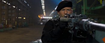 Movie still from “Expend4bles” (2023), directed by Scott Waugh – A man in a black jacket and a black hat is holding a rifle; Medium shot, Low angle
