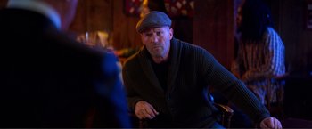 Movie still from “Expend4bles” (2023), directed by Scott Waugh – A man wearing a hat and a sweater is sitting down; Medium shot, Over the shoulder angle