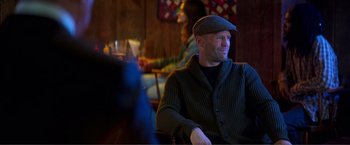 Movie still from “Expend4bles” (2023), directed by Scott Waugh – A man sitting in front of other people in a room; Medium shot, Over the shoulder angle