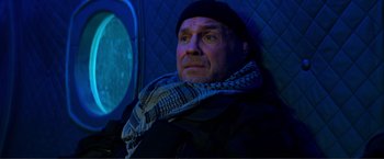Movie still from “Expend4bles” (2023), directed by Scott Waugh – A man wearing a scarf and a hat is sitting on a plane; Medium shot, Low angle