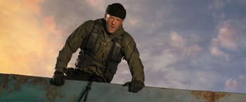 Movie still from “Expend4bles” (2023), directed by Scott Waugh – A man standing on top of a building holding a snowboard; Medium shot, Low angle