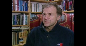 Movie still from “Explorer” (2022), directed by Matthew Dyas – A person wearing a jacket in front of a book shelf; Close Up shot, High angle