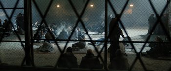 Movie still from “Extraction II” (2023), directed by Sam Hargrave – A group of people sitting on the ground behind a chain link fence; Wide shot, High angle
