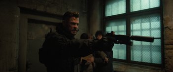 Movie still from “Extraction II” (2023), directed by Sam Hargrave – A man holding a gun in a room with other people; Medium shot, Over the shoulder angle