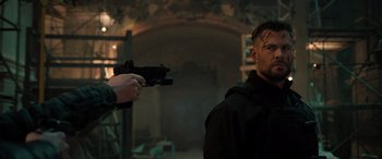 Movie still from “Extraction II” (2023), directed by Sam Hargrave – A man holding a gun pointed at another man in a dark room; Medium shot, Over the shoulder angle