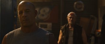 Movie still from “F9: The Fast Saga” (2021), directed by Justin Lin – A man standing next to another man in a room; Close Up shot, Over the shoulder angle