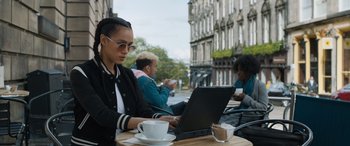 Movie still from “F9: The Fast Saga” (2021), directed by Justin Lin – A woman sitting at an outdoor table using a laptop computer; Medium shot, Over the shoulder angle