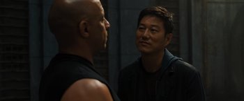 Movie still from “F9: The Fast Saga” (2021), directed by Justin Lin – A man standing next to another man in front of a brick wall; Close Up shot, Over the shoulder angle