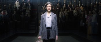 Movie still from “Fantastic Beasts and Where to Find Them” (2016), directed by David Yates – A woman in a gray jacket holding a purse in front of a group of people; Medium shot, High angle