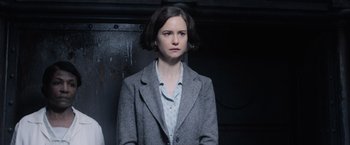 Movie still from “Fantastic Beasts and Where to Find Them” (2016), directed by David Yates – A woman in a gray coat standing next to a wall; Medium shot, Over the shoulder angle