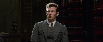 Movie still from “Fantastic Beasts: The Crimes of Grindelwald” (2018), directed by David Yates – A man wearing a suit and tie in front of a book case; Close Up shot, Low angle