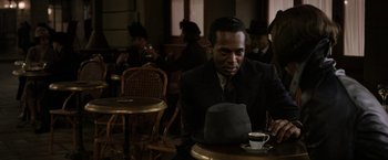 Movie still from “Fantastic Beasts: The Crimes of Grindelwald” (2018), directed by David Yates – A man sitting at a table with a hat on his head; Medium shot, Over the shoulder angle