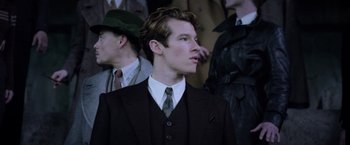 Movie still from “Fantastic Beasts: The Crimes of Grindelwald” (2018), directed by David Yates – A man in a suit and tie standing in front of a group of people; Close Up shot, Over the shoulder angle