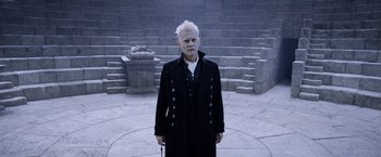 Movie still from “Fantastic Beasts: The Crimes of Grindelwald” (2018), directed by David Yates – A man in a black coat stands in front of a stone wall; Medium shot, High angle