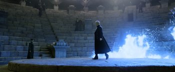 Movie still from “Fantastic Beasts: The Crimes of Grindelwald” (2018), directed by David Yates – A man in a black coat walking in front of a stone wall; Wide shot, High angle