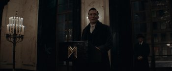 Movie still from “Fantastic Beasts: The Secrets of Dumbledore” (2022), directed by David Yates – A man standing in front of a podium holding a laptop; Medium shot, Low angle